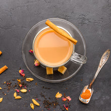 Load image into Gallery viewer, Image of a glass tea cup and saucer containing masala chai. Surrounded by fresh spices and a metal spoon with some chai masala