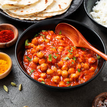 Load image into Gallery viewer, Image of a ceramic bowl containing chickpea curry and a wooden spoon. Surrounded by small bowls of turmeric and chilli powder, as well as a partial view of a plate of naan bread and rice