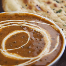 Load image into Gallery viewer, Image of a Urad Dal/Dal Makhni with a swirl of cream on the top. With a partial view of a naan bread